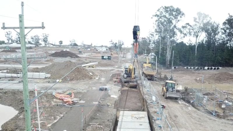 Aerial drone view down a box culvert trench with a crane hook, excavators and trucks working along the site