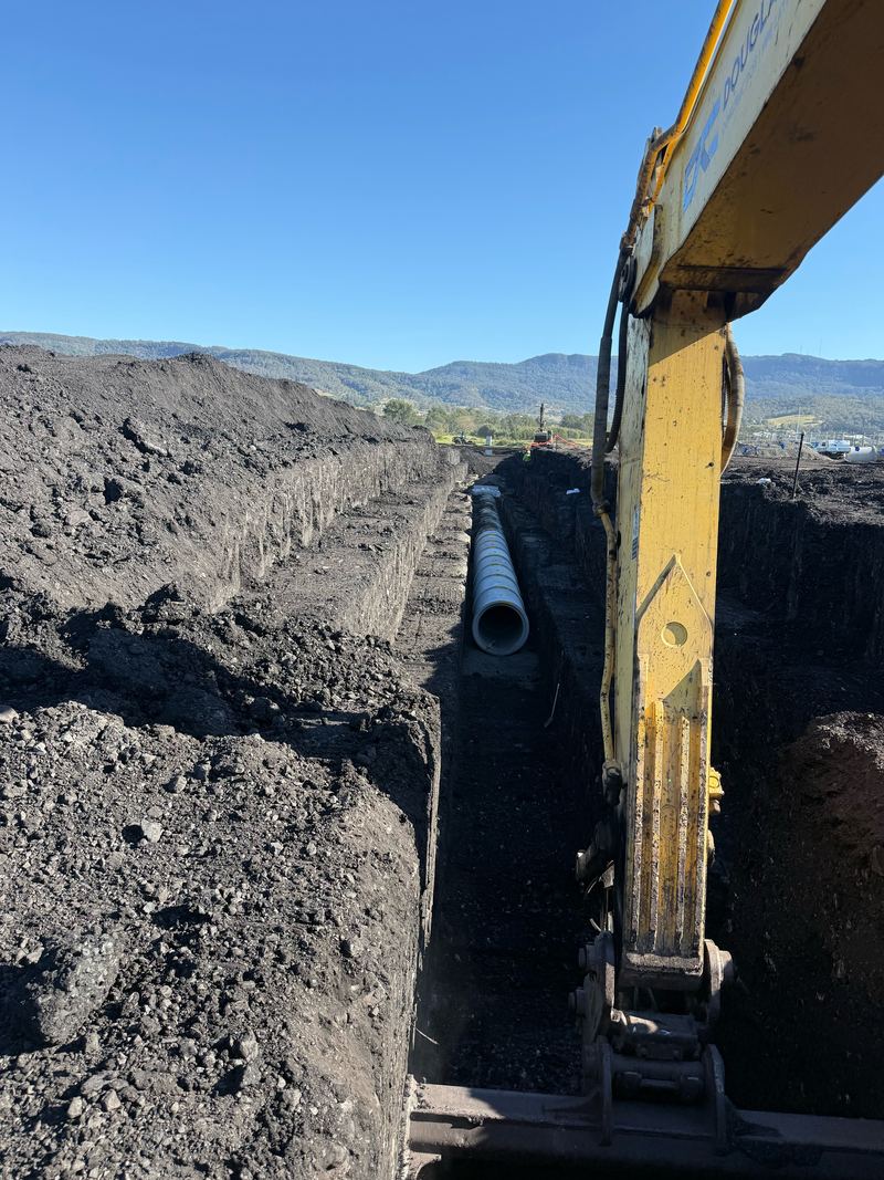 Stormwater pipe going into a deep benched trench, Douglass Civil branded excavator arm in the foreground