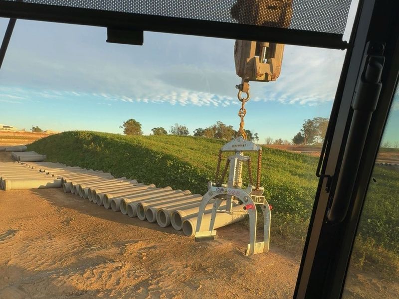 View from an excavator cab of a pipe lifter attachment with concrete pipes lined up, late afternoon